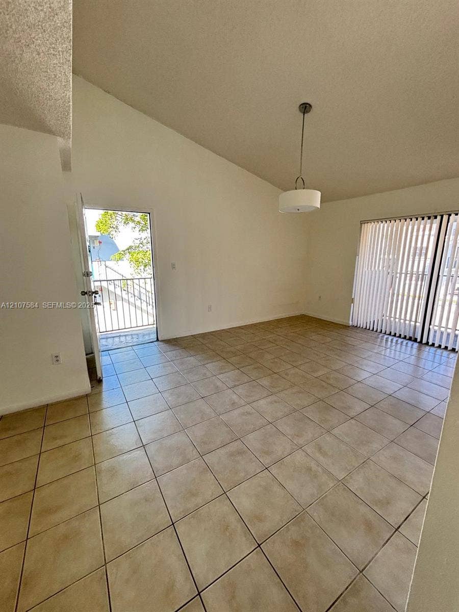 Vaulted ceiling, neutral tile flooring, drum-style pendant lighting, and sliding glass doors with vertical blinds