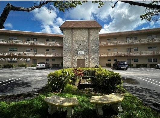 5003 NW 35th Street, #506, Lauderdale Lakes, FL 33319 Multi-story facade featuring a textured stone central column, shingle roofing, and decorative railings on the upper levels, complemented by a landscaped grounds area with dual stone benches