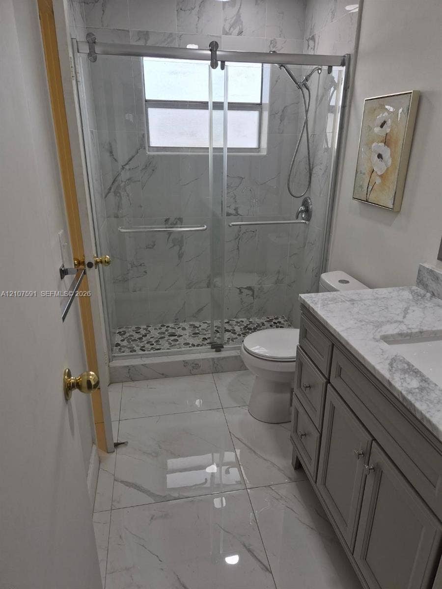 5003 NW 35th Street, #506, Lauderdale Lakes, FL 33319 Bathroom featuring a gray vanity with stone-style countertop, shower enclosure with sliding glass doors, stone-style wall and floor tiling, and a built-in pebble-style shower floor