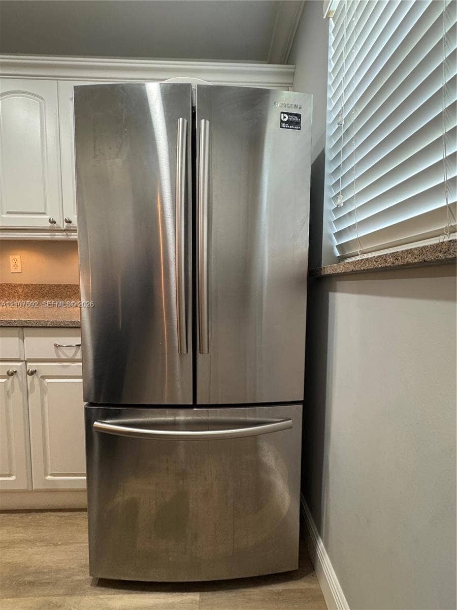 Stainless steel French door refrigerator featuring a bottom-mount freezer, integrated white cabinetry, stone-surface countertops, and horizontal blind window treatment