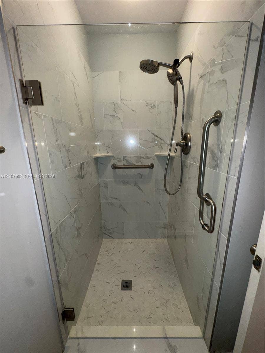 Walk-in shower featuring large-format tile surround with a marble-finish aesthetic, frameless glass enclosure, adjustable-height handheld showerhead, and integrated metal grab bars