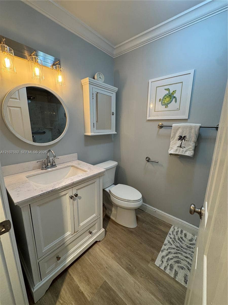 White vanity with a stone countertop, oval mirror, wall-mounted medicine cabinet, wood-finish flooring, and crown molding