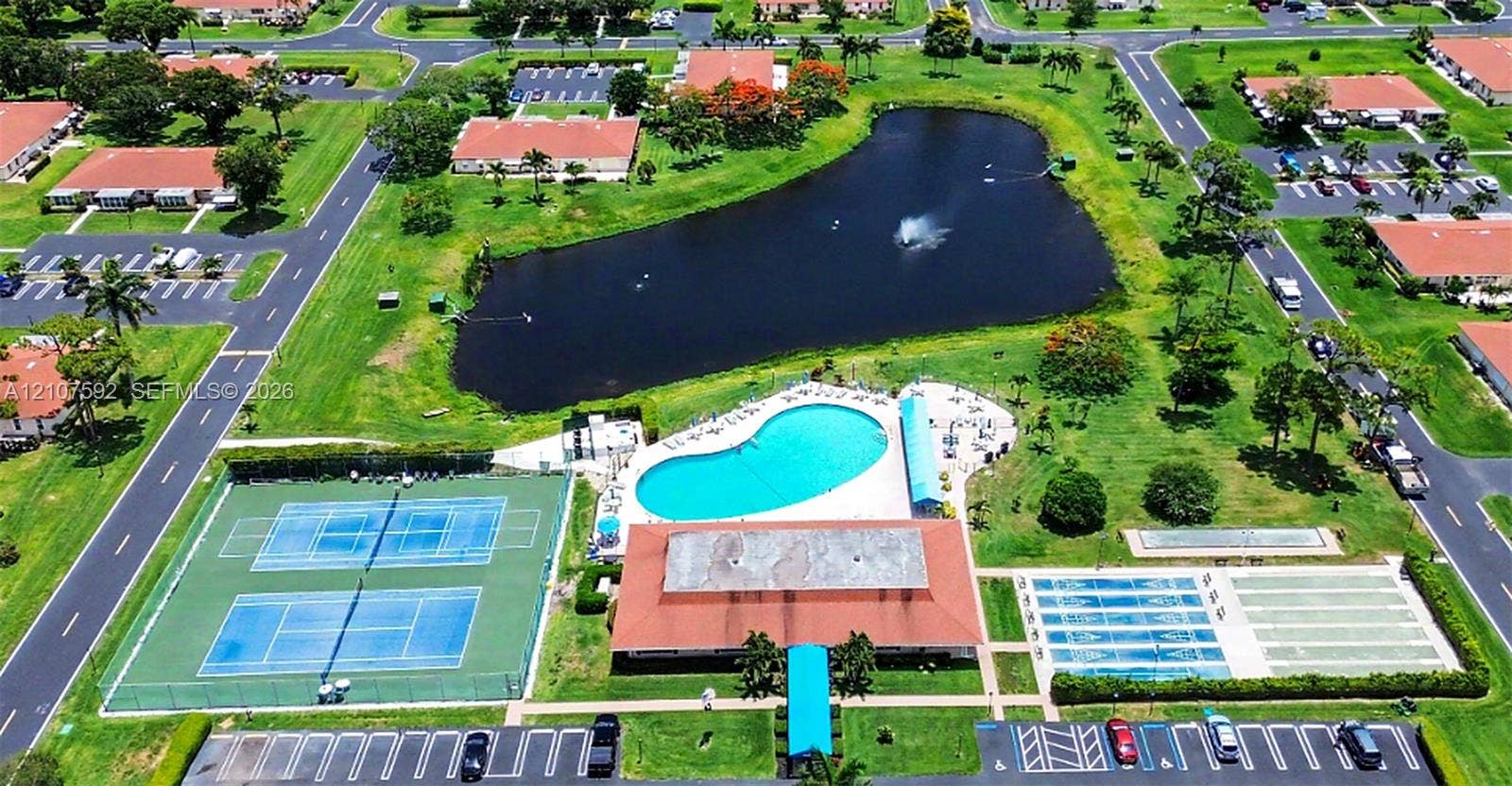 Aerial perspective of community recreational facilities featuring a central lake with fountain, swimming pool, two hard-court tennis courts, and multi-use athletic courts