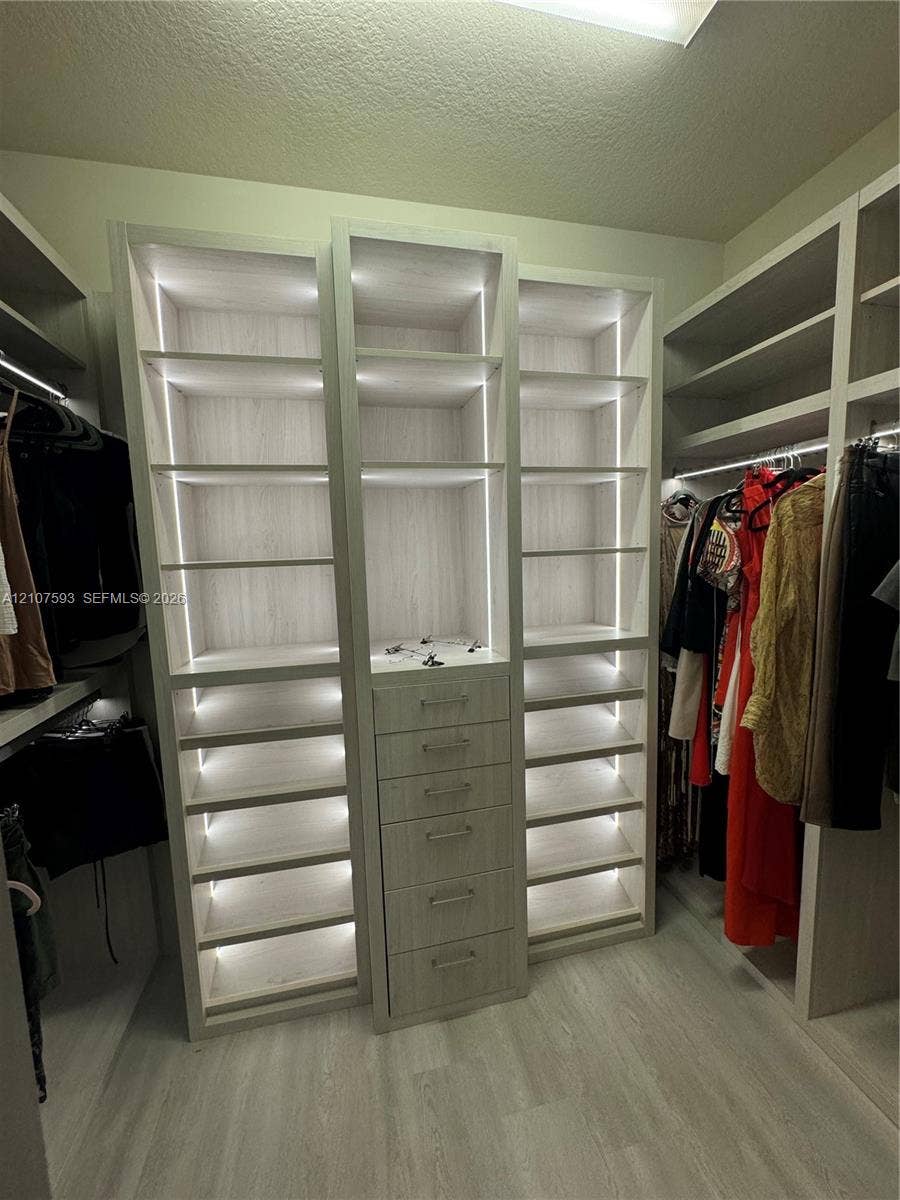 Walk-in closet featuring custom light-toned cabinetry, integrated vertical accent lighting, five stacked storage drawers, and wood-finish flooring
