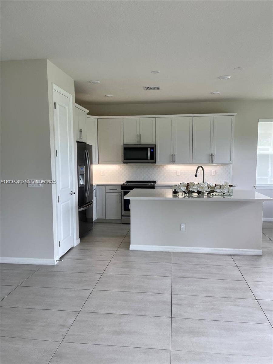 Kitchen featuring white shaker-style cabinetry, a central island with a solid-surface countertop, stainless steel appliances, under-cabinet lighting, and large-format tile flooring