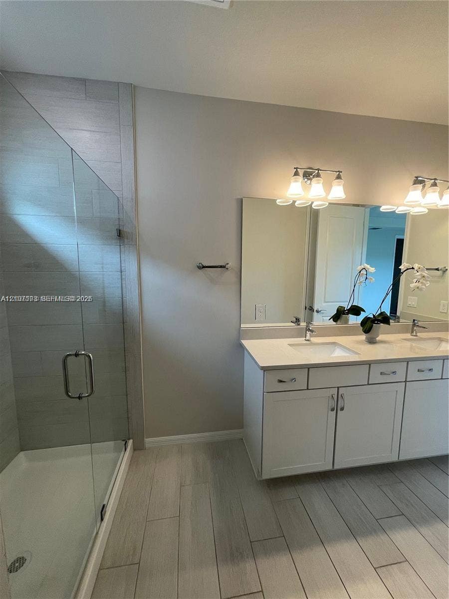Double vanity with white cabinetry and solid-surface countertops, walk-in shower with floor-to-ceiling tile surround and glass enclosure, and wood-finish tile flooring