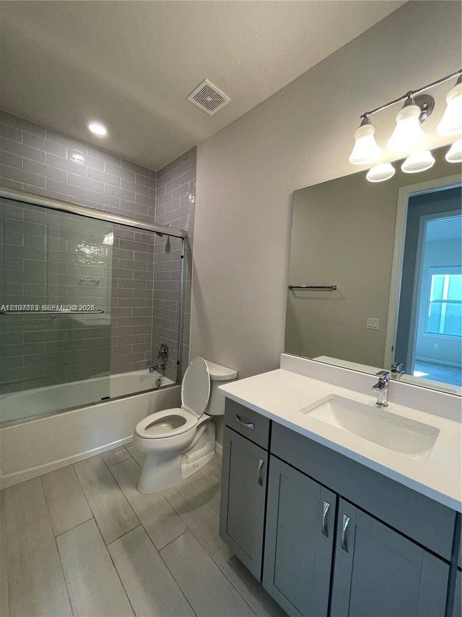 Bathroom featuring a gray vanity with a stone countertop, an integrated rectangular sink, wood-finish flooring, and a bathtub with a gray subway tile surround and sliding glass door
