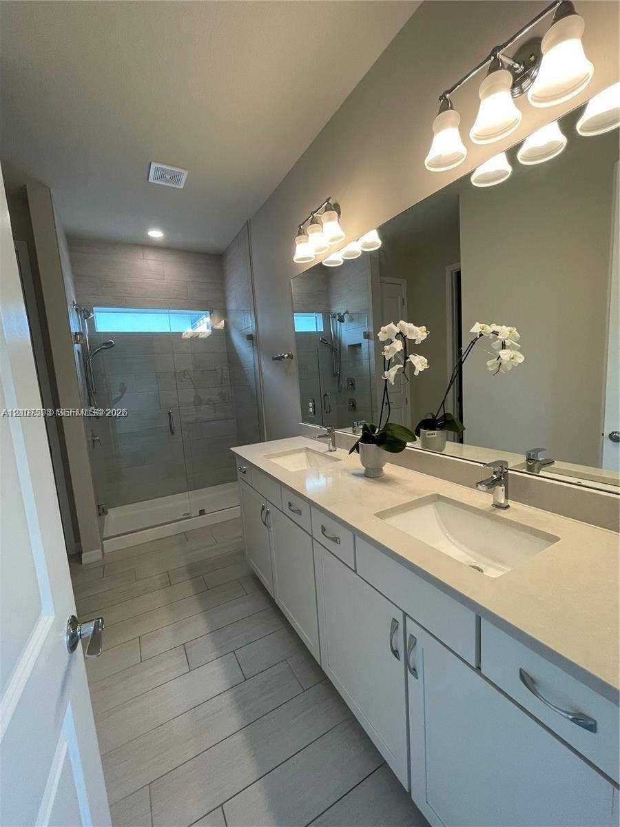 Dual vanity featuring rectangular undermount sinks, quartz countertop, cabinetry with silver hardware, full-width mirror, and a glass-enclosed shower with tiled wall surrounds