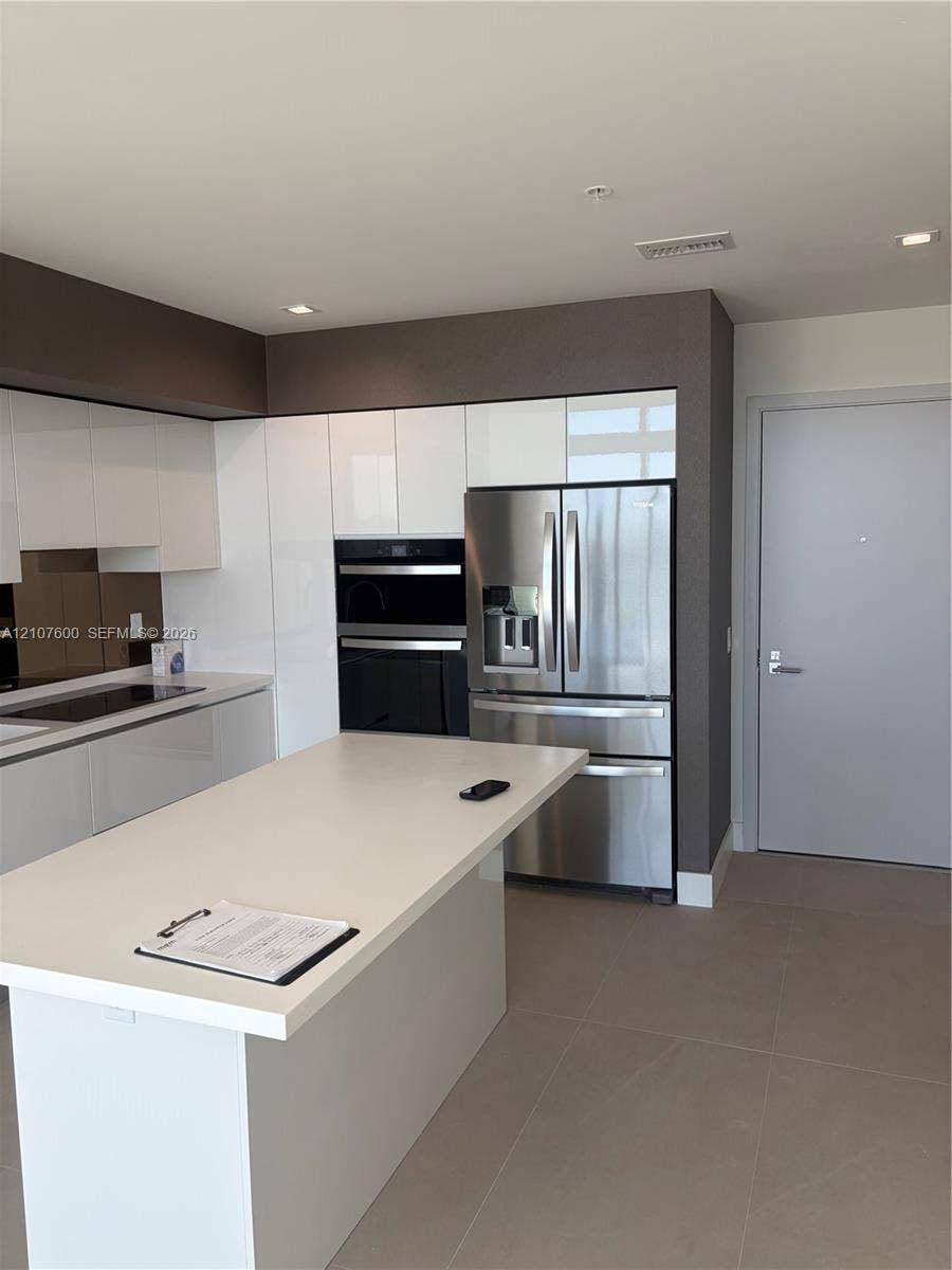Kitchen featuring a central island with a solid surface countertop, high-gloss white cabinetry, stainless steel appliances including a built-in wall oven, and ceramic tile flooring