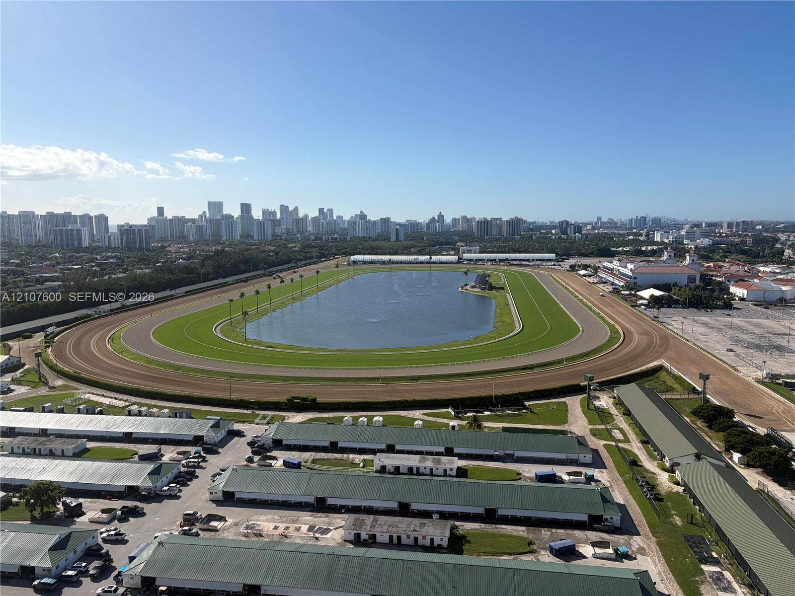 Aerial perspective of a large-scale track facility featuring a central grassed oval, an expansive interior water feature, a multi-lane dirt track, and rows of metal-roofed structures