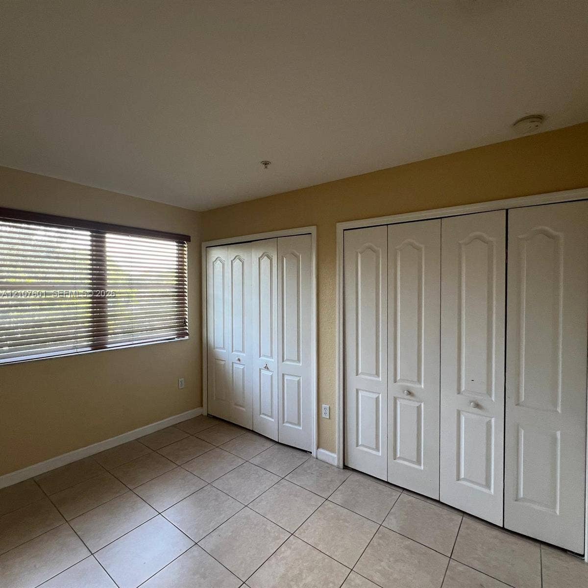 7080 NW 177th Street, #203-13, Hialeah, FL 33015 Interior room featuring tile flooring, two sets of white paneled bi-fold closet doors, and a window equipped with horizontal blinds