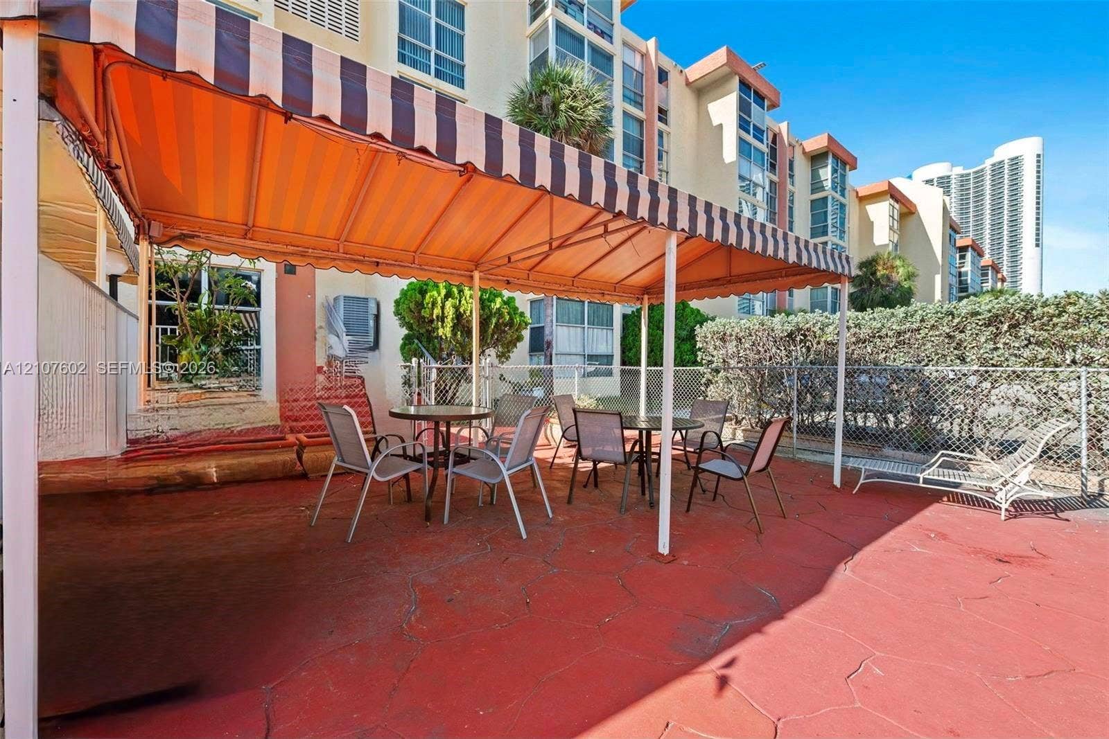 210 172nd Street, #542, Sunny Isles Beach, FL 33160 Ground-level patio featuring red-toned concrete flooring, a retractable striped fabric awning, and perimeter chain-link fencing