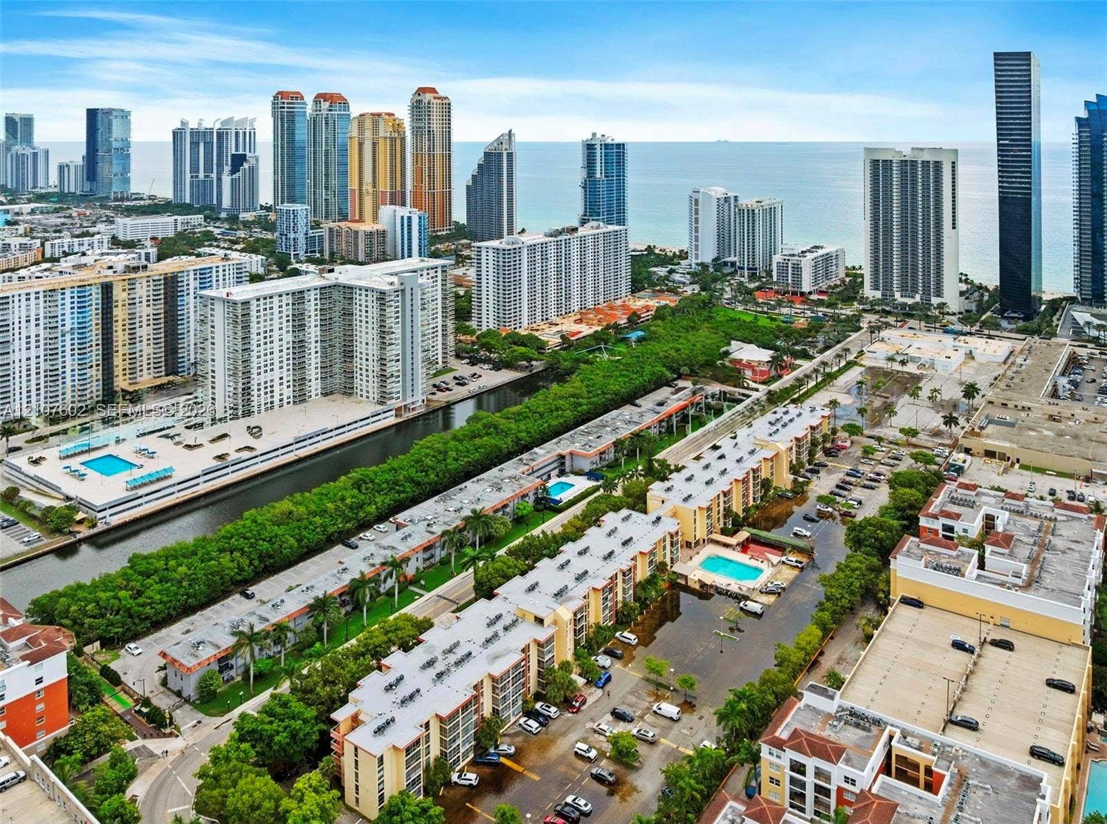 210 172nd Street, #542, Sunny Isles Beach, FL 33160 Aerial perspective featuring multiple mid-rise residential structures, surface parking areas, landscaped grounds, and proximity to an urban coastline