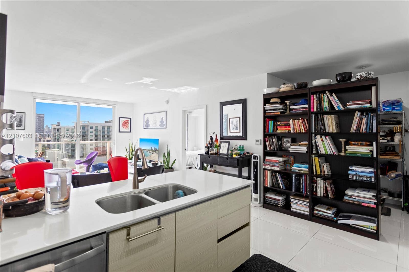 4250 Biscayne Boulevard, #1418, Miami, FL 33137 Kitchen island featuring a white countertop, stainless steel dual-basin sink, and light-tone cabinetry with horizontal grain, complemented by white polished floor tiling and floor-to-ceiling glass sliding doors