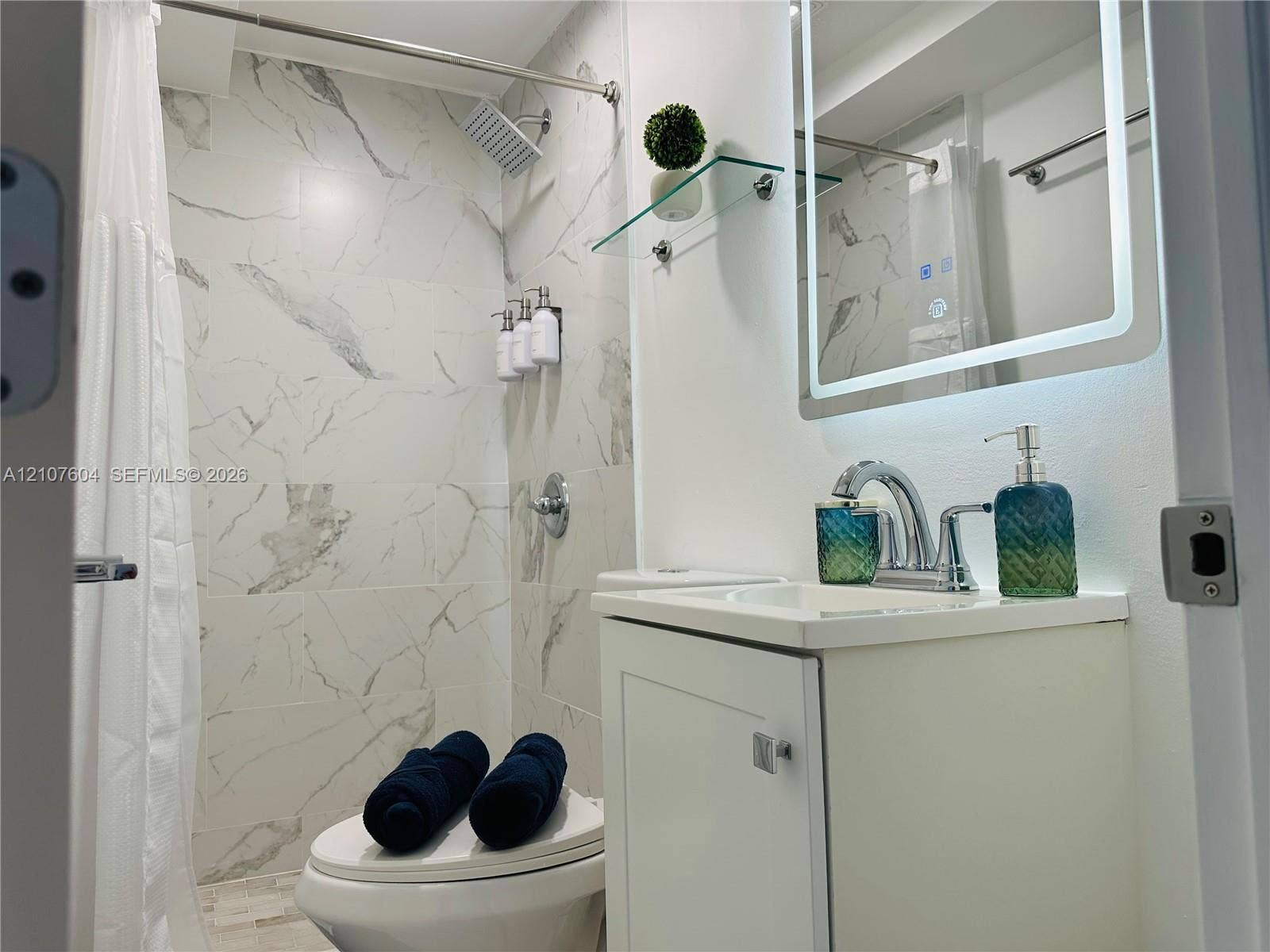 120 S 56th Avenue, Hollywood, Hollywood, FL 33023 Bathroom featuring a shower stall with stone-finish tile surround, a chrome rain-style shower head, a white vanity with an integrated sink, a chrome widespread faucet, and a wall-mounted LED-backlit mirror