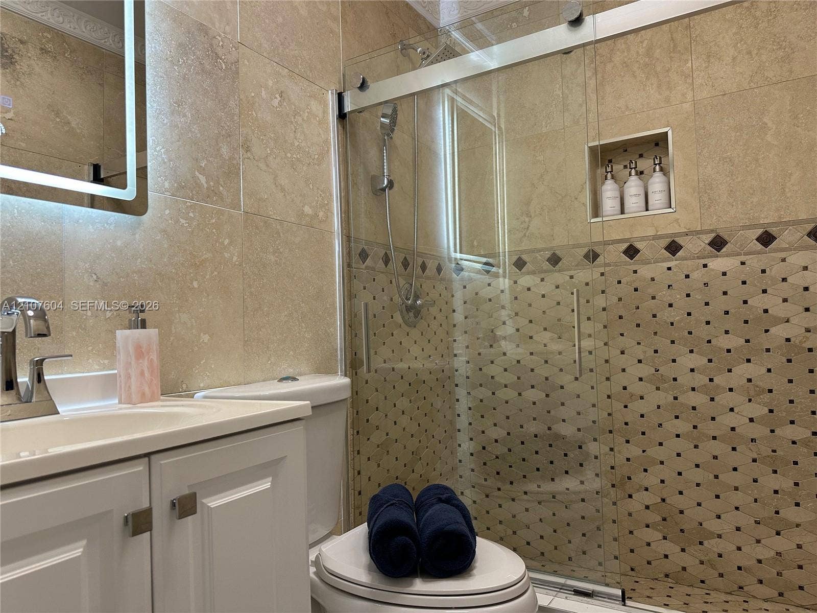 120 S 56th Avenue, Hollywood, Hollywood, FL 33023 Bathroom featuring a vanity with a white cabinet, stone-tile wall surfacing, a walk-in shower with a glass sliding door, a recessed shower niche, and a wall-mounted illuminated mirror