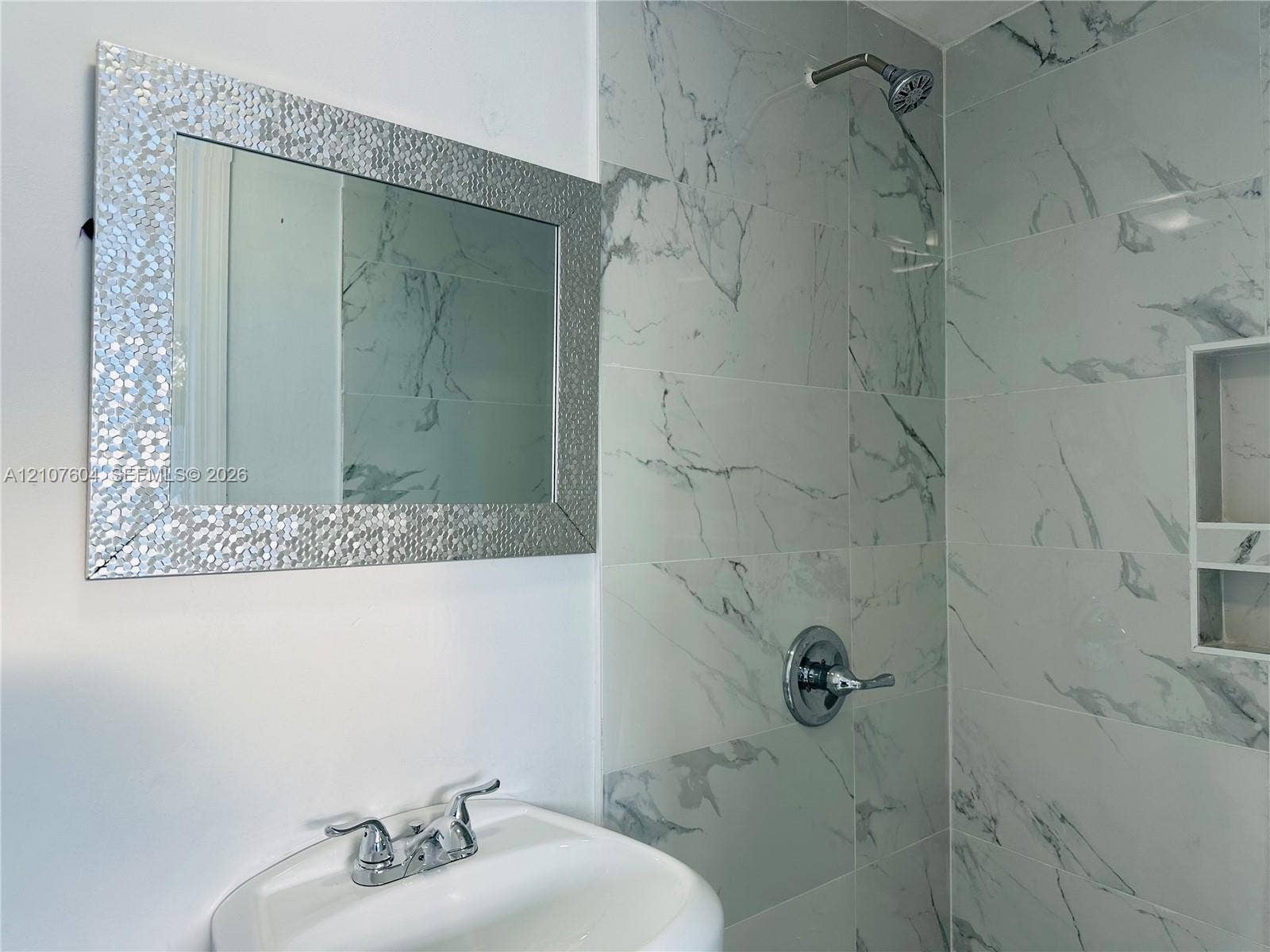 120 S 56th Avenue, Hollywood, Hollywood, FL 33023 Wall-mounted bathroom vanity featuring a basin sink with a two-handle chrome faucet, accompanied by a rectangular decorative-frame mirror and a walk-in shower with large-format stone-patterned tile, chrome hardware, and an integrated recessed soap niche