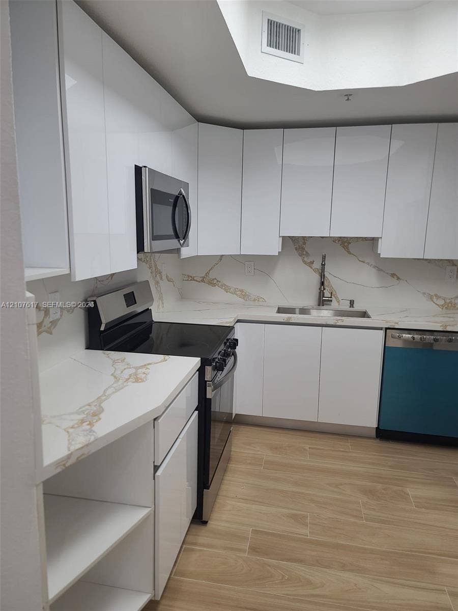 17415 NW 75th Place, #103, Hialeah, Hialeah, FL 33015 Modern kitchen featuring high-gloss white cabinetry, stone-finish countertops with matching backsplash, stainless steel appliances, wood-finish flooring, and a recessed ceiling skylight