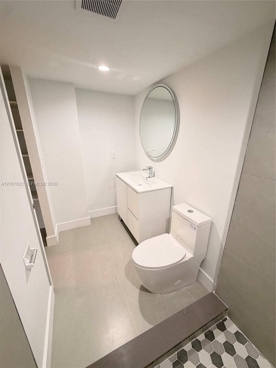 20400 W Country Club Drive, #112, Aventura, FL 33180 Bathroom featuring a white vanity cabinet, rectangular integrated sink, oval wall-mounted mirror, dual-flush toilet, and hexagonal floor tile