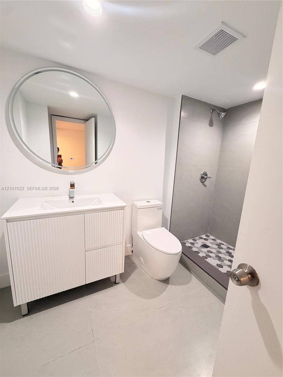 20400 W Country Club Drive, #112, Aventura, FL 33180 Bathroom featuring a white fluted vanity with a rectangular sink, a large circular mirror, a dual-flush toilet, and a walk-in shower with large-format grey wall tile and geometric mosaic floor tile