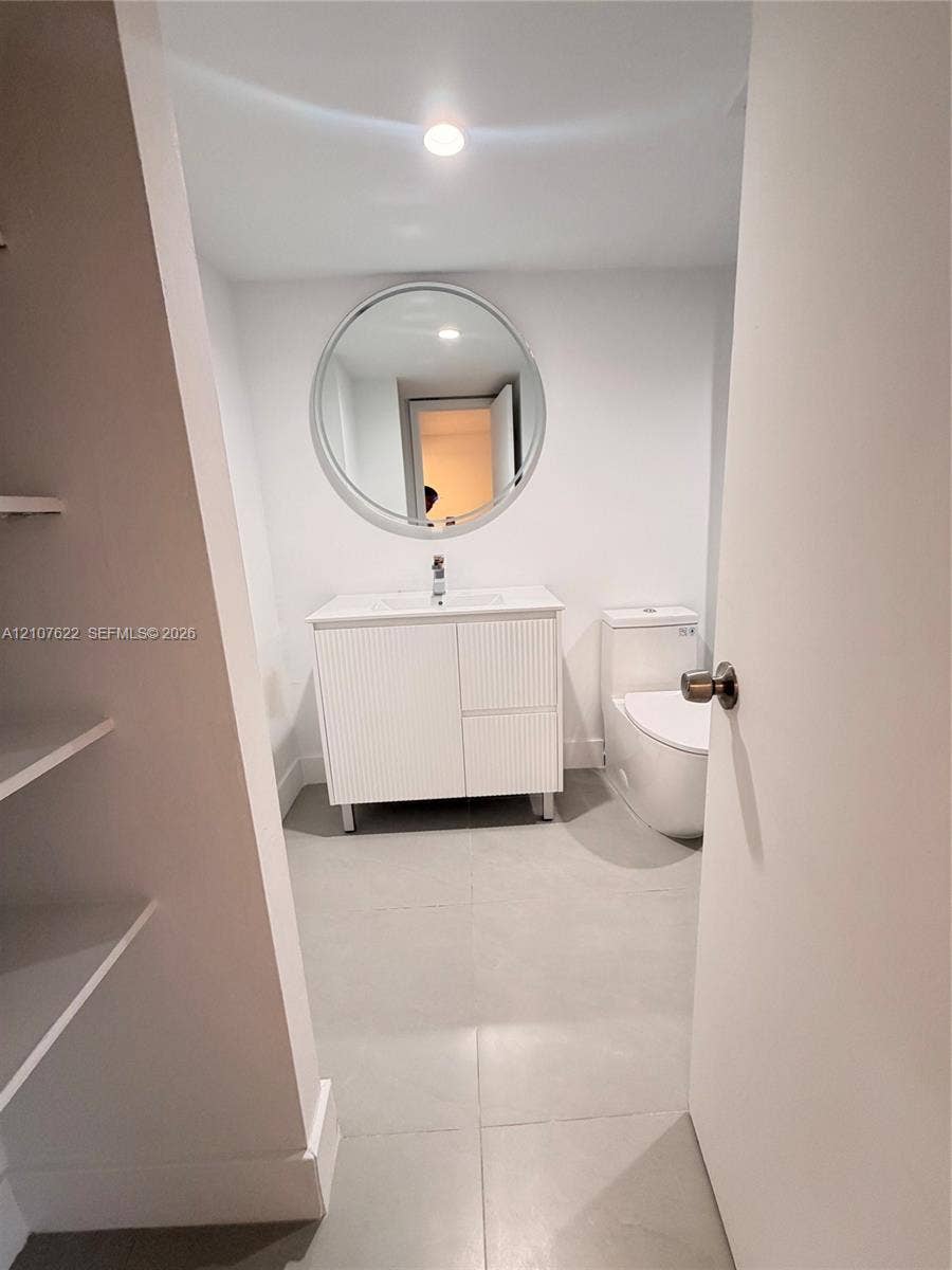 20400 W Country Club Drive, #112, Aventura, FL 33180 Bathroom featuring a ribbed-texture vanity, round mirror, white countertop with integrated sink, large-format tile flooring, and a skirted toilet