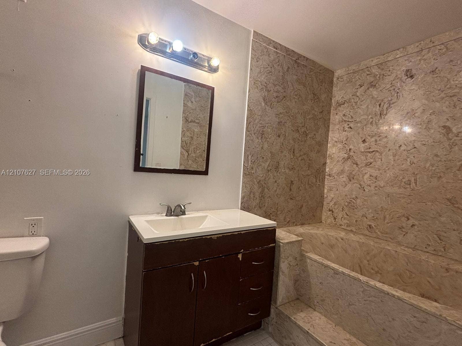 19 Crossings Circle, #H, Boynton Beach, Boynton Beach, FL 33435 Bathroom featuring a vanity with an integrated sink, three-light wall-mounted fixture, framed mirror, and a tub surround with stone-patterned paneling