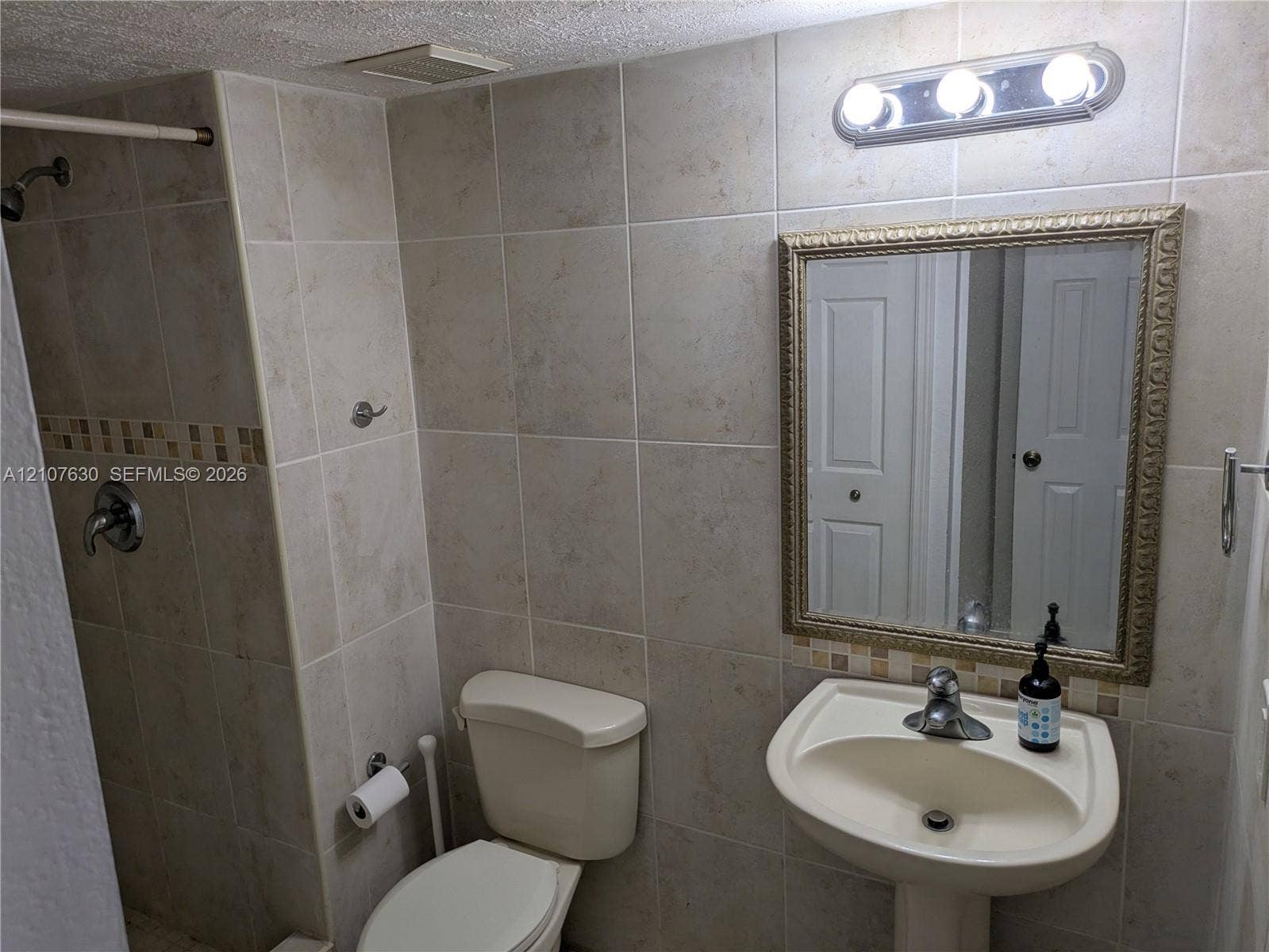 Bathroom featuring beige wall tiles with a glass mosaic accent band, a white pedestal sink with a chrome faucet, a framed wall mirror, and a walk-in shower