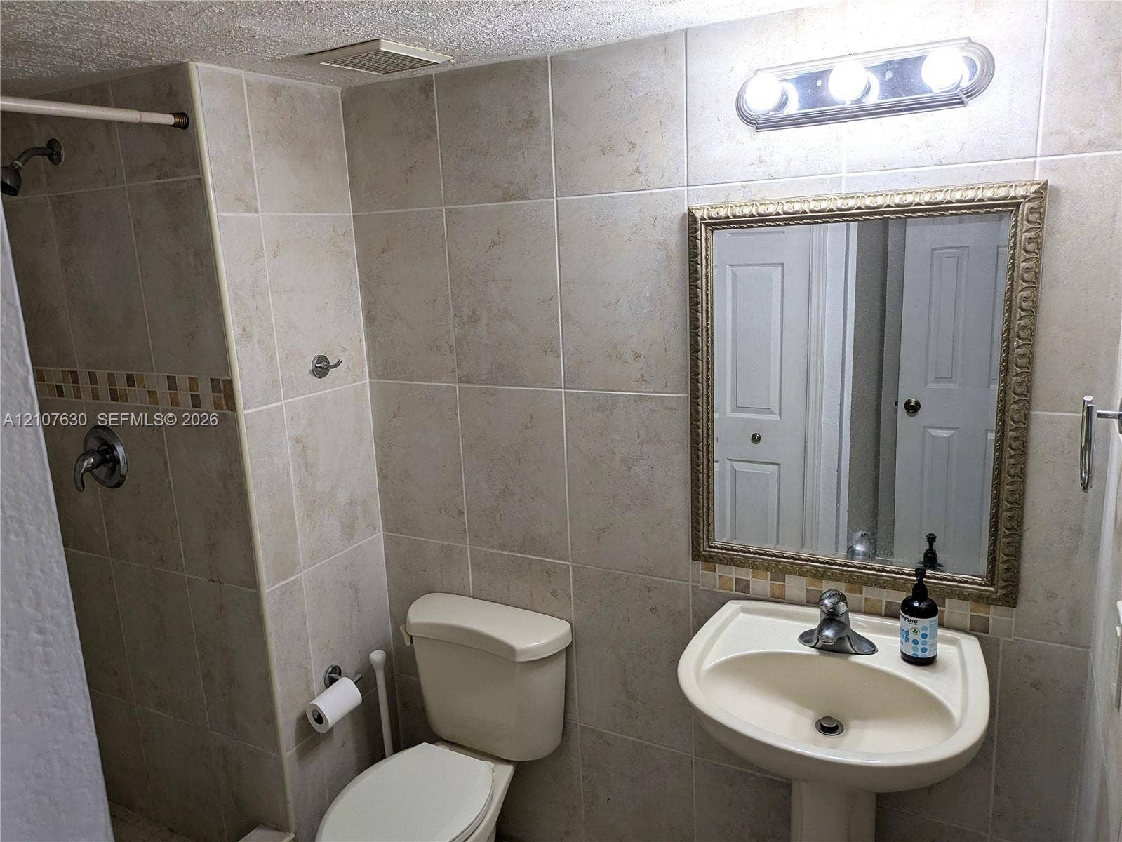 Full-height wall tiling with decorative mosaic inlay, pedestal sink with chrome faucet, and glass-front medicine cabinet with integrated light fixture