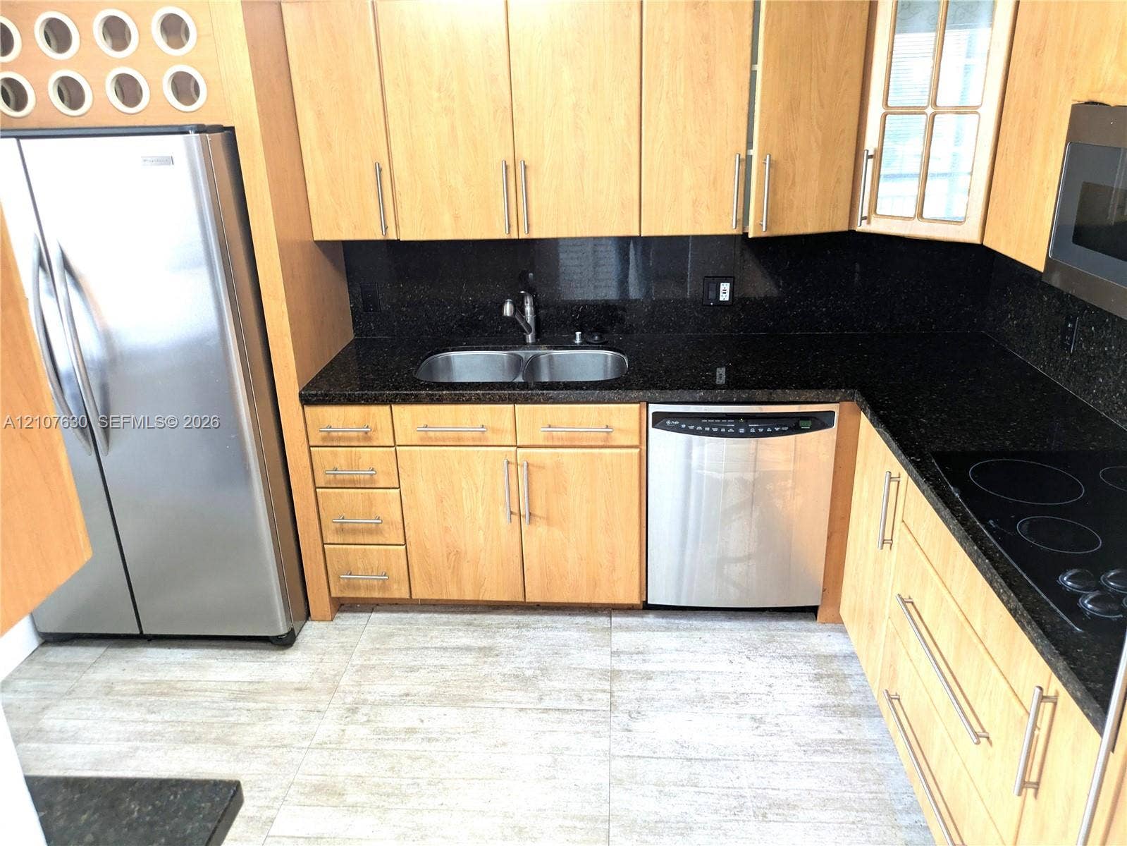 Kitchen featuring light-finish cabinetry, dark solid-surface countertops, dual-basin stainless steel sink, tile flooring, and integrated stainless steel appliances