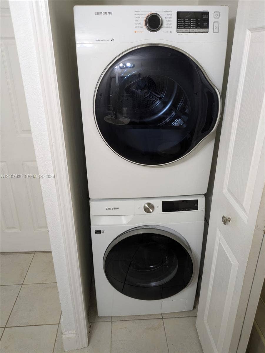 Stacked white front-load washer and dryer units with digital controls and glass-paneled doors, situated within a recessed laundry closet featuring tile flooring