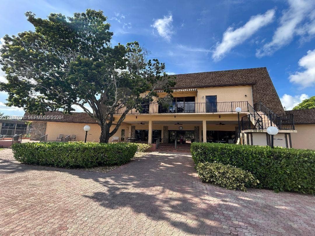151 Berkley Road, #112, Hollywood, FL 33024 Multi-story building featuring an expansive brick paver entrance, a wraparound second-level balcony with metal railing, and mature landscaping