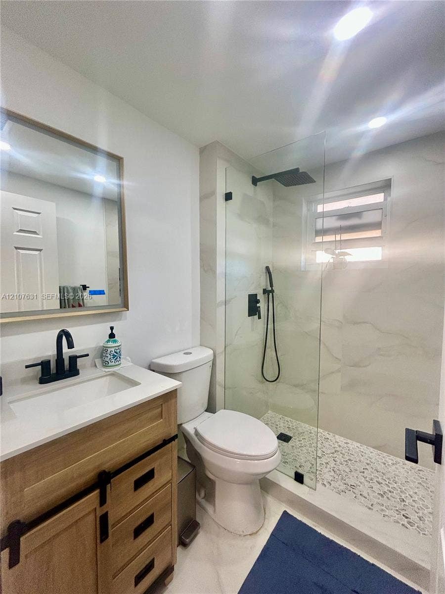 151 Berkley Road, #112, Hollywood, FL 33024 Wood-finish vanity with a light-tone countertop, frameless glass-enclosed shower featuring large-format tile walls, pebble-stone shower flooring, and matte black plumbing fixtures