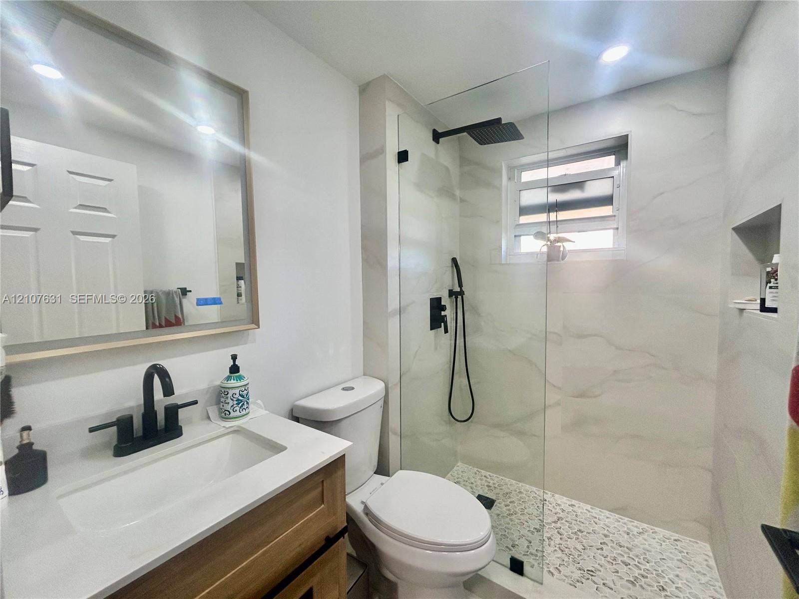 151 Berkley Road, #112, Hollywood, FL 33024 Bathroom featuring a wood-finish vanity with a white countertop, matte black fixtures, a walk-in shower with a glass panel, and a built-in recessed wall niche