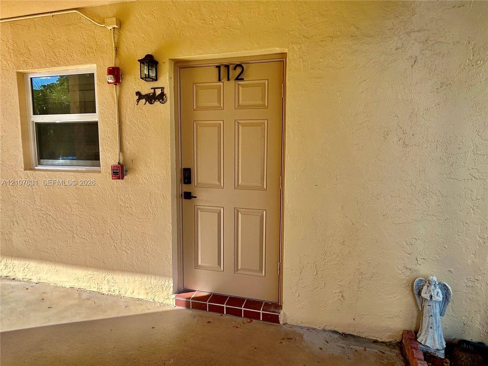 151 Berkley Road, #112, Hollywood, FL 33024 Stucco exterior featuring a six-panel entry door, single-hung window with a metallic frame, and exterior wall-mounted carriage-style lighting