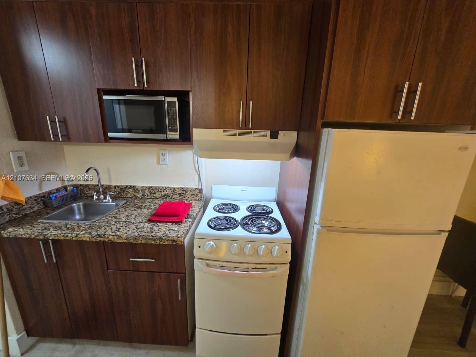 19201 Collins Avenue, #941, Sunny Isles Beach, FL 33160 Kitchenette featuring dark wood-finish cabinetry, stone-patterned countertops, stainless steel sink with high-arch faucet, built-in microwave, and electric range