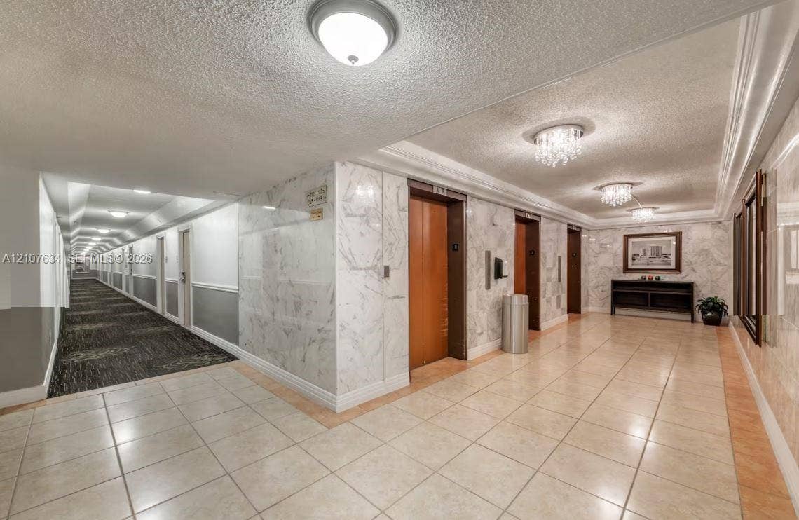 19201 Collins Avenue, #941, Sunny Isles Beach, FL 33160 Common area lobby featuring natural stone wall cladding, polished tile flooring, multiple elevator banks with wood-finish doors, and decorative crystal-style ceiling flush-mount lighting