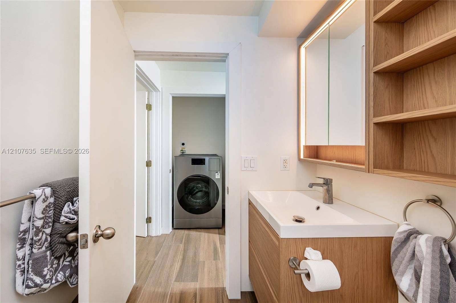 217 Madeira Avenue, #3, Coral Gables, Coral Gables, FL 33134 Bathroom featuring a floating vanity with a light wood finish, integrated white basin, illuminated medicine cabinet, built-in open shelving, and stainless steel washer and dryer