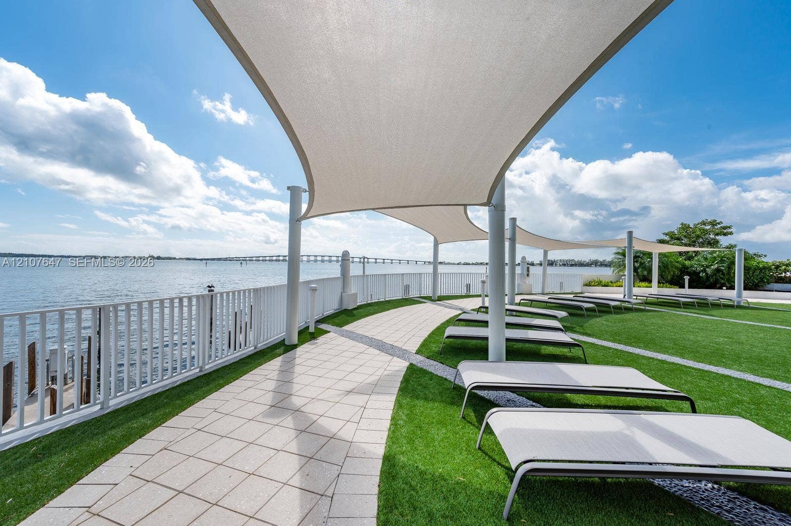 200 SE 15th Road, #PH-A, Miami, Miami, FL 33129 Waterfront patio featuring expansive white sail shade structures, synthetic turf lawn, stone-paver walkway, and white metal railing