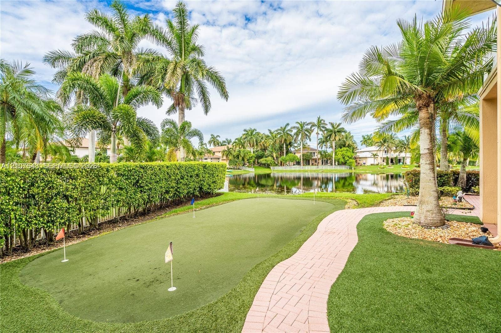 Backyard featuring a synthetic putting green, brick paver walkway, mature palm trees, privacy hedging, and water frontage