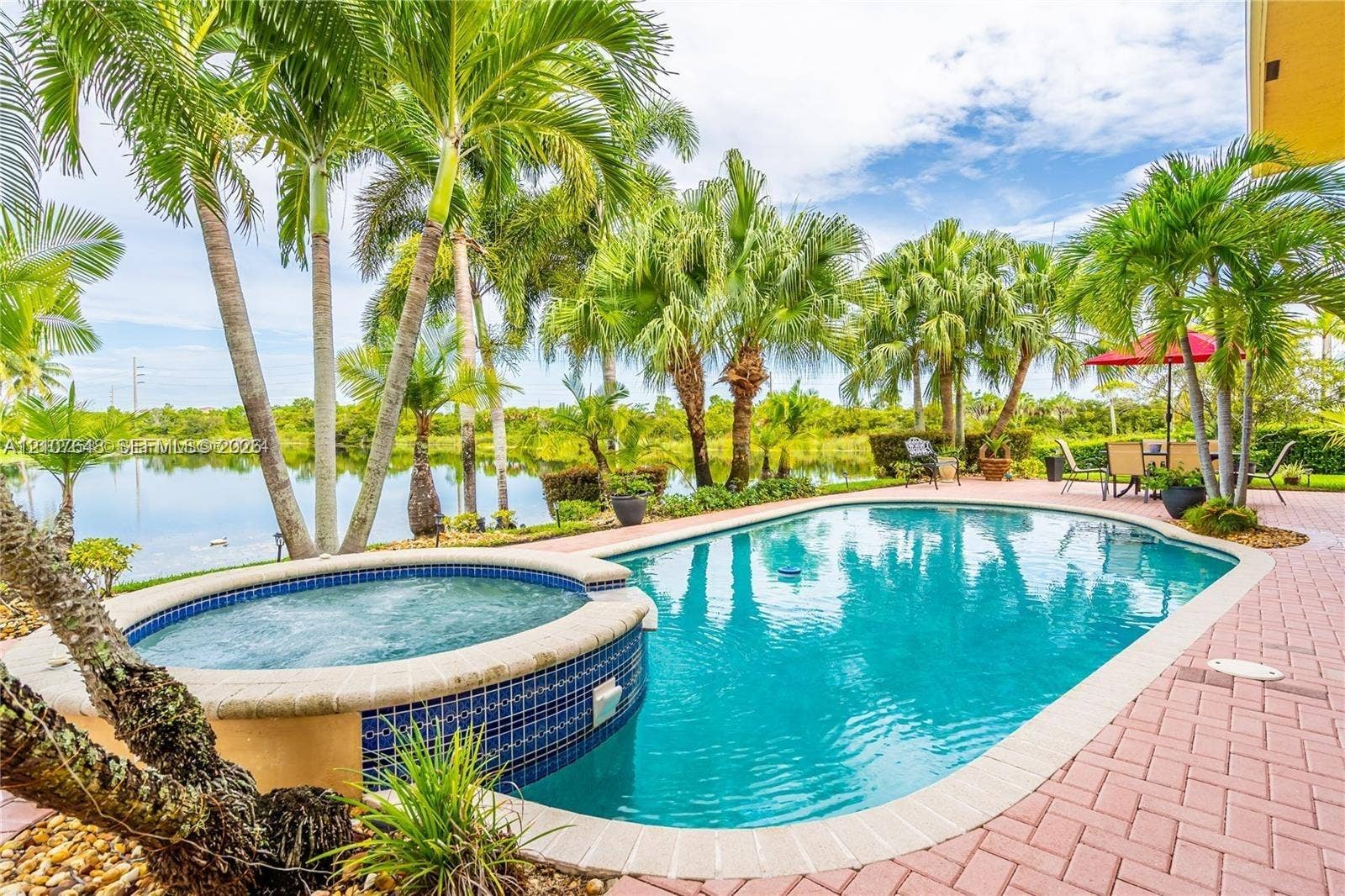 Inground swimming pool with an attached spa, finished with blue glass tile and a light-toned coping, surrounded by a brick-paver patio and mature palm trees overlooking a pond