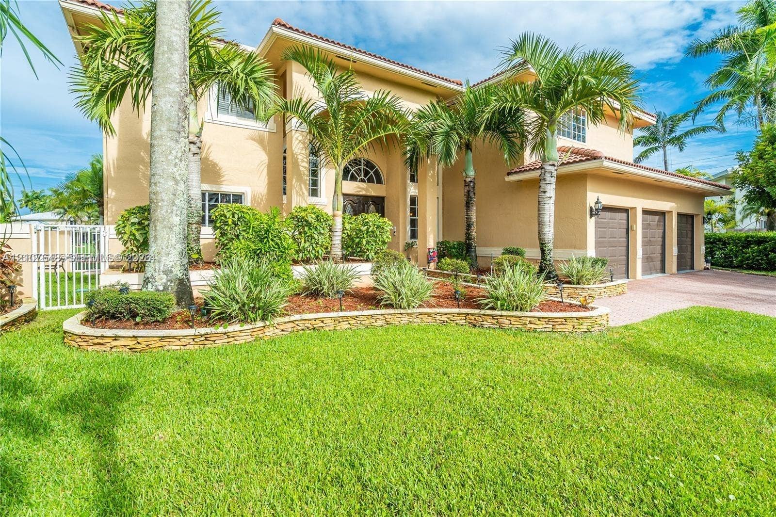 Two-story residence featuring stucco exterior, multi-car garage, arched entryway, and tiered stone garden beds with mature palm trees