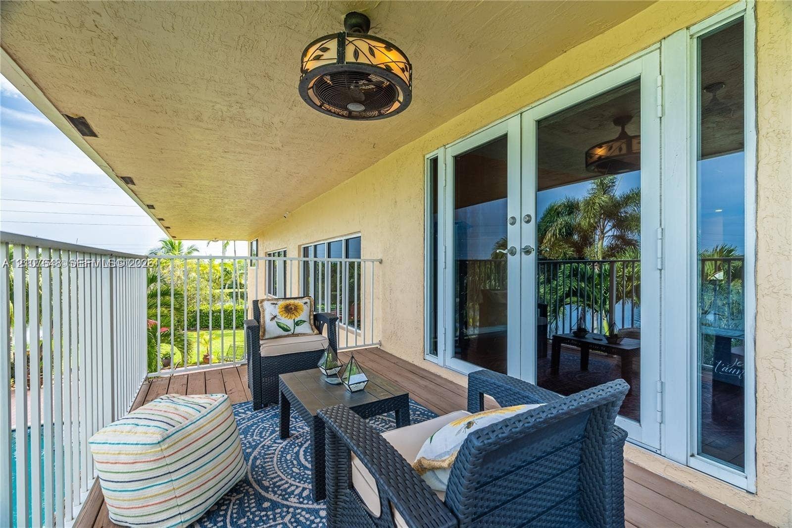 Covered balcony featuring wood-finish flooring, white metal railing, and a decorative ceiling-mounted fan with integrated lighting, accessed by full-length glass paneled double doors
