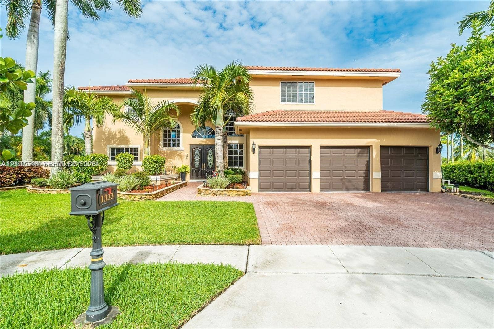 Two-story residence featuring a terracotta tile roof, stucco exterior, three-car garage, paver driveway, and arched entryway with decorative glass double doors