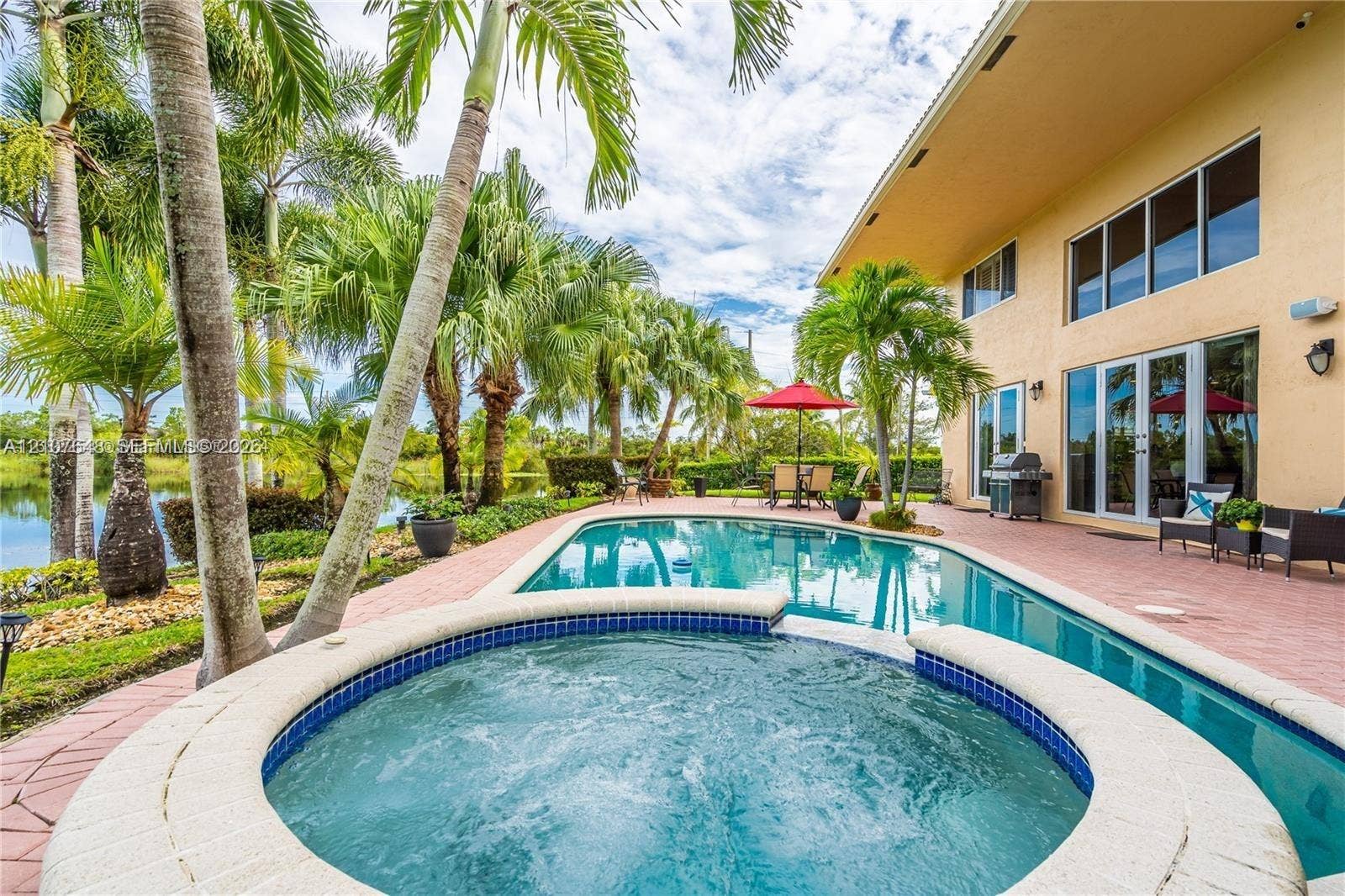 In-ground swimming pool and spa with blue tile accents, surrounded by a brick-paver patio, mature palm trees, and waterfront perimeter