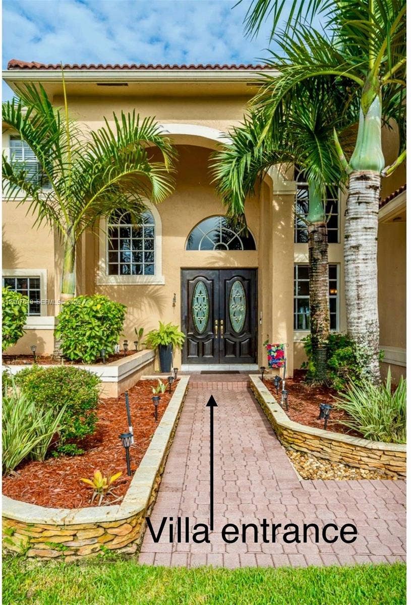 Stucco exterior featuring an arched entryway with double doors, decorative glass insets, transom window, and stone-accented raised landscape beds along a paved walkway