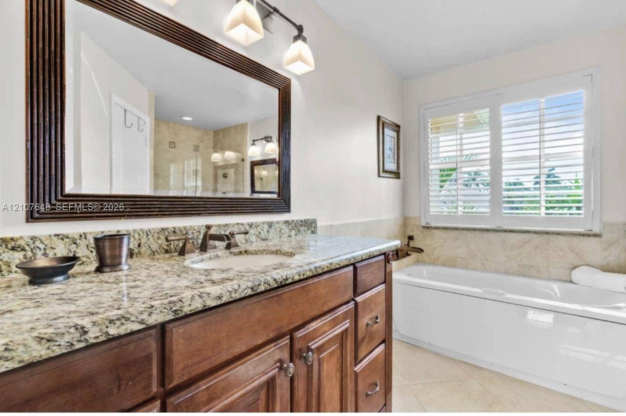 Wood-finish vanity cabinet, stone countertop with integrated sink, rectangular framed mirror with integrated lighting, soaking tub with tile surround, and plantation shutters