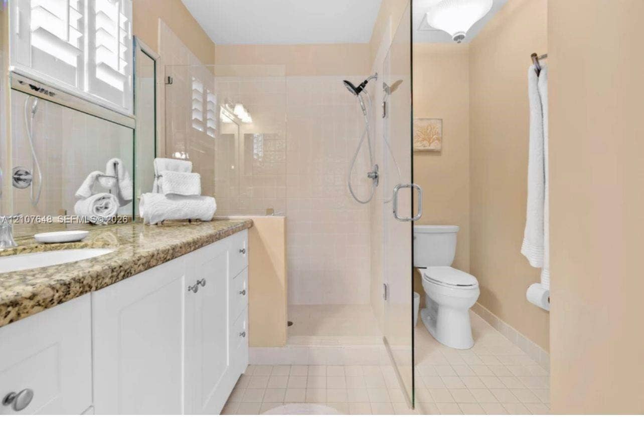 Bathroom featuring a white vanity cabinet with stone-look countertop, an integrated sink, a glass-enclosed walk-in shower with a handheld showerhead, and shutter window coverings
