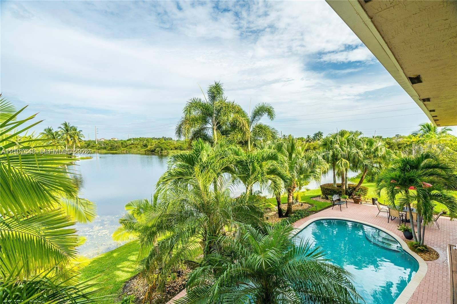 Outdoor living area featuring a paved brick patio, a curved swimming pool, mature palm trees, and waterfront grounds