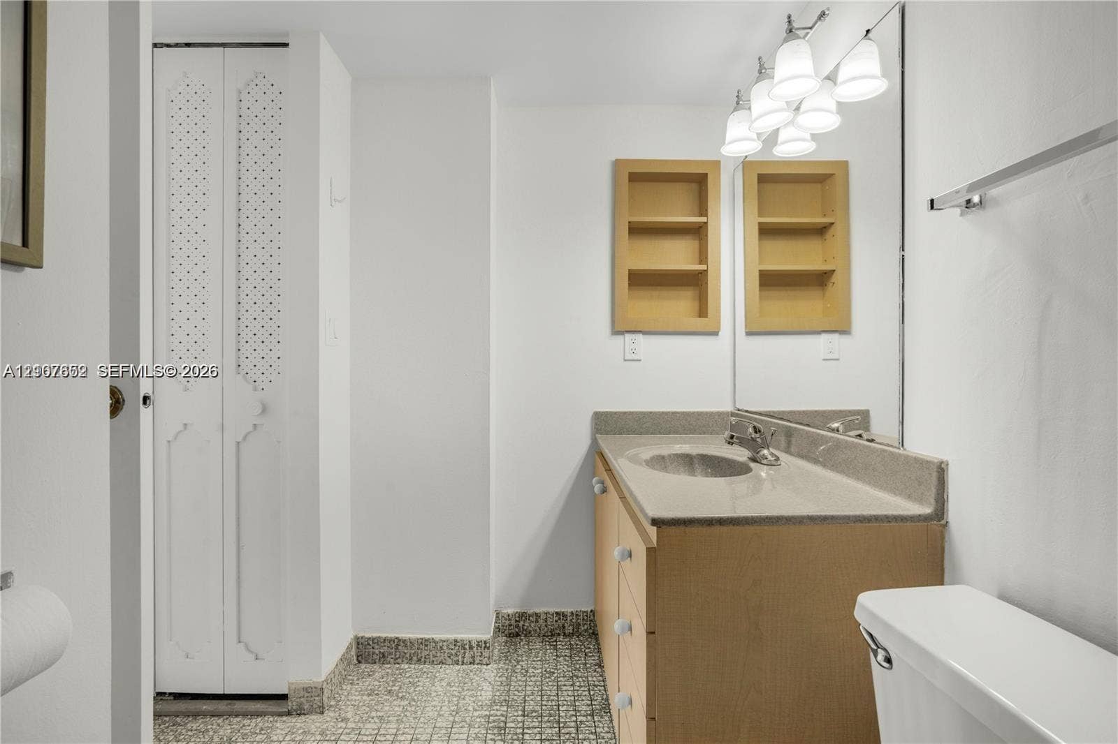 20500 W Country Club Drive, #607, Aventura, Aventura, FL 33180 Bathroom vanity with a solid-surface countertop and integrated sink, recessed wall shelving, multi-light fixture, full-width mirror, and bi-fold louvered closet doors