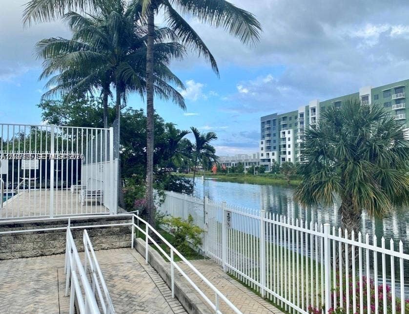 2088 NE 167th Street, #2-111, North Miami Beach, North Miami Beach, FL 33162 Waterfront outdoor area featuring concrete paver decking, white metal fencing, mature palm trees, and a tiered retaining wall